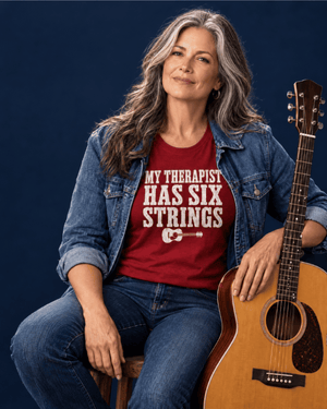 A woman with gray hair sits on a stool holding an acoustic guitar, wearing a red graphic t-shirt that reads 'MY THERAPIST HAS SIX STRINGS' with a white guitar illustration, layered under a blue denim jacket against a dark blue background.