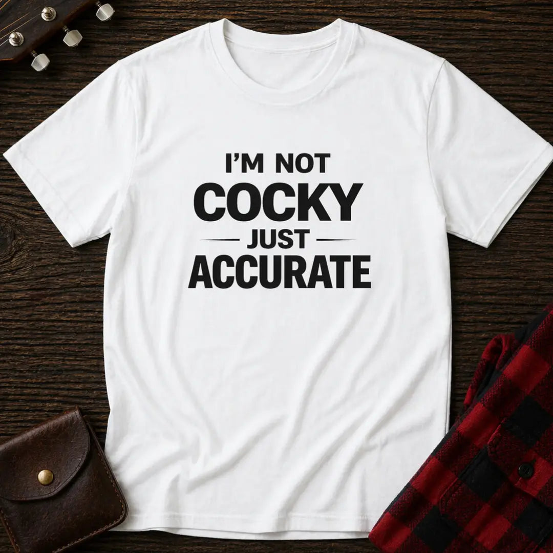 White t-shirt with 'I'm not cocky just accurate' text on a wooden surface.