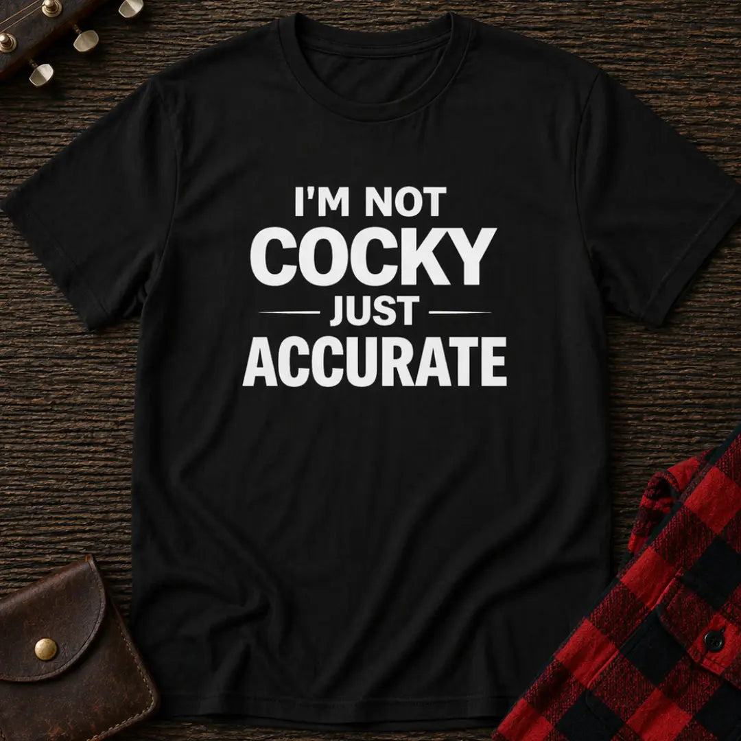 Black t-shirt with 'I'm not cocky just accurate' text on a wooden surface with a plaid shirt and leather item.