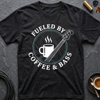A black short-sleeve graphic T-shirt laid flat on a dark textured surface, featuring a circular white and teal print that reads “FUELED BY COFFEE & BASS” around an illustration of a coffee mug and a bass guitar. Small styled props including a guitar pick, gold-toned accessory, coffee cup, and cable are visible around the shirt.