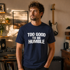 Man wearing a navy blue t-shirt with 'Too Good To Be Humble' text in a room with a guitar and decor.